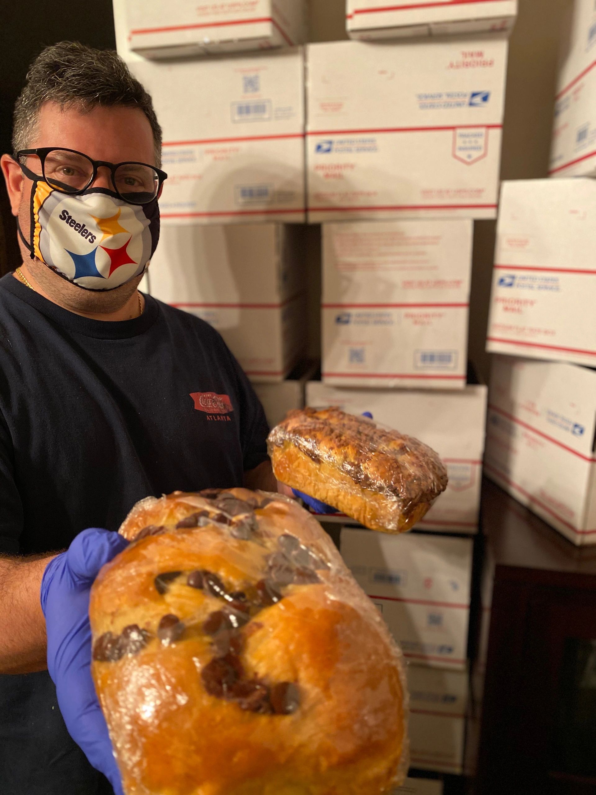 Man wearing Steelers mask and gloves holding wrapped baked goods in front of packed USPS boxes.