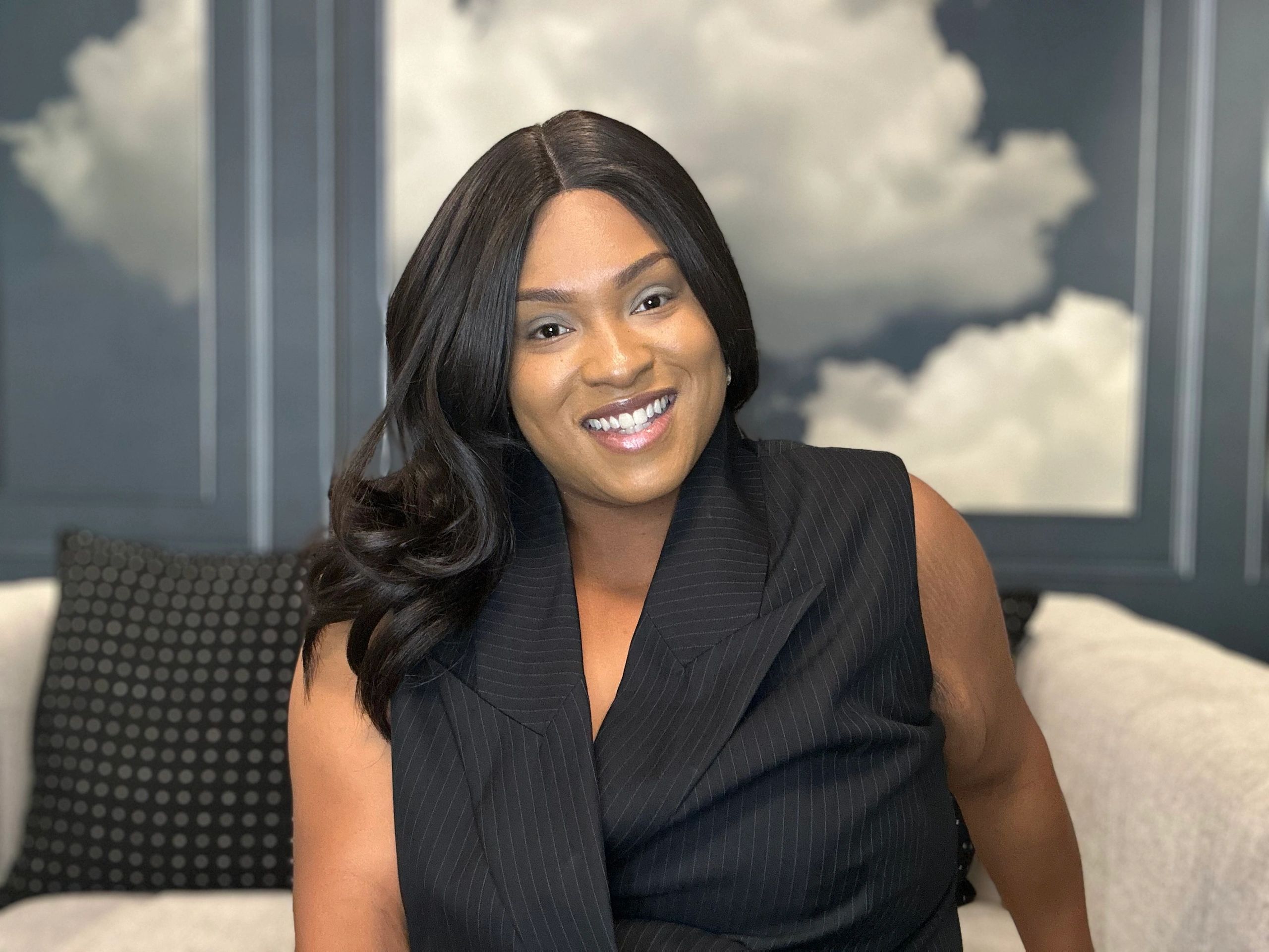 A smiling woman in a black outfit sitting on a couch with a cloudy sky background.
