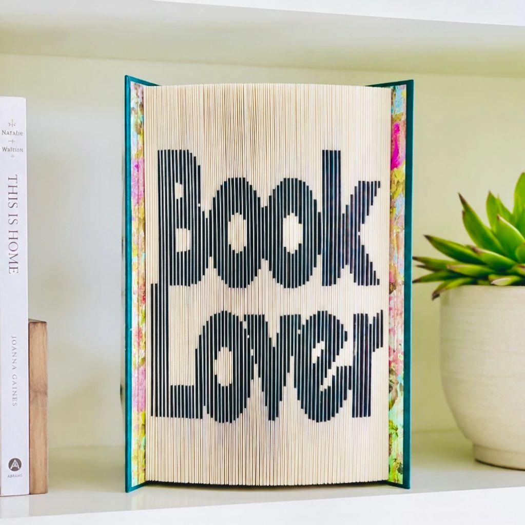 Book art with pages cut out to spell Book Lover, stacked on top of each other. Set on shelf w plant.