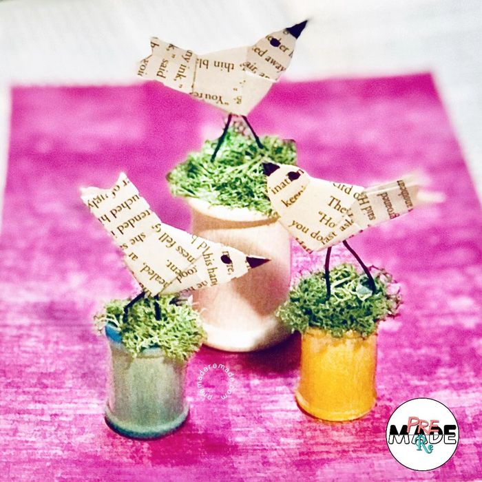paper birds created from book pages, set atop small empty thread spools topped with green moss 