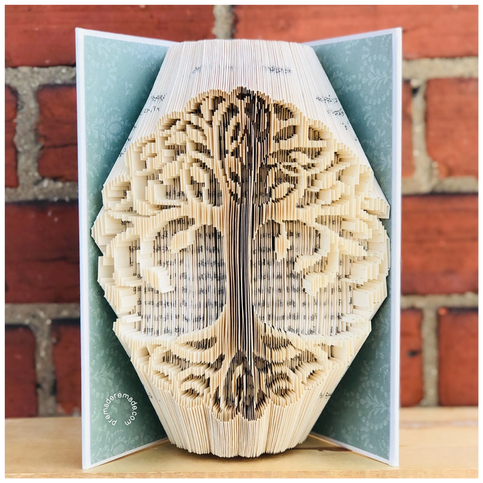 white hardcover book with a detailed tree carved into it's pages, with a brick wall behind it