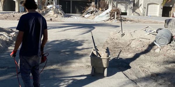 A man stands near a wheelbarrow in a dusty, debris-filled street.