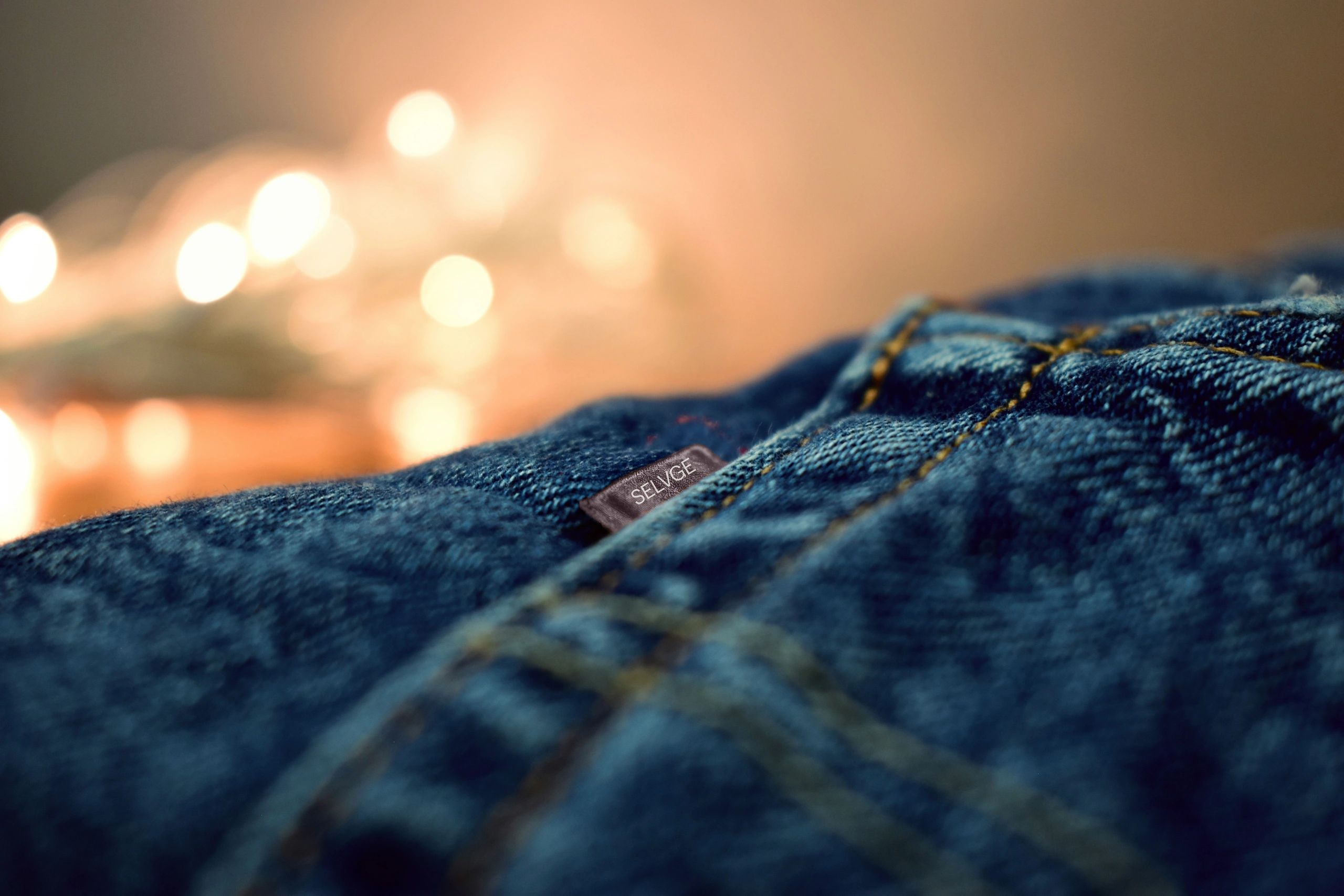Close-up of denim jeans with a selvedge tag and blurred lights in background.