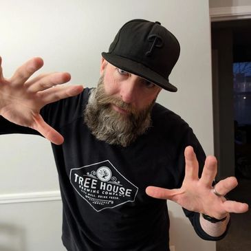 Bearded man in a black cap and Tree House Brewing shirt posing with hands outstretched.