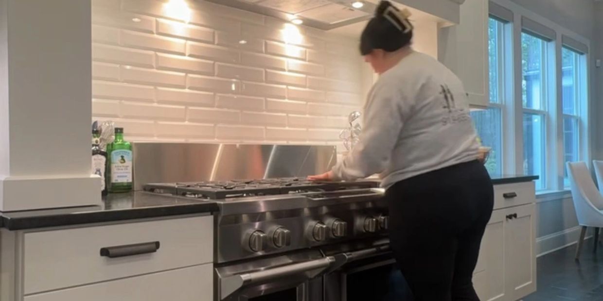 A woman cleans a modern kitchen stove with white cabinets and large windows.