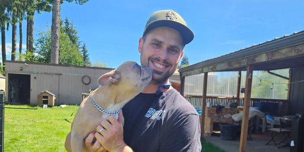 Man holding and being licked by a dog outdoors.