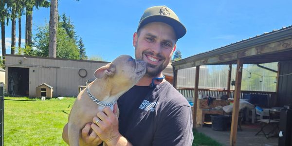 Man smiling while holding a dog that licks his face.