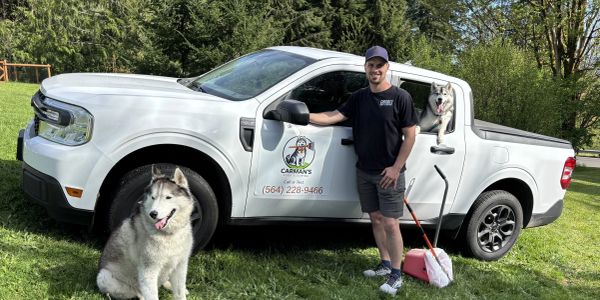 Man with two huskies near a white pickup truck on grass.