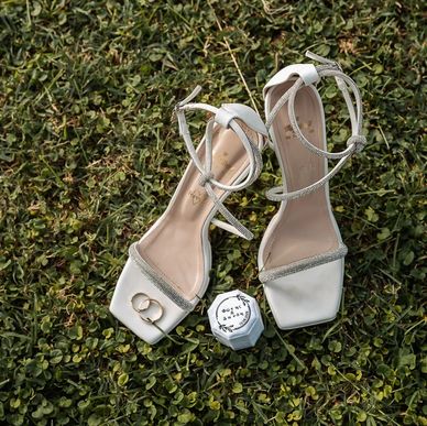 Elegant white heels with wedding rings on grass.