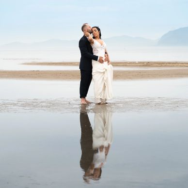 A couple in wedding attire stands barefoot on a reflective beach.