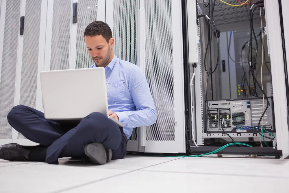Man working on laptop inside a server room, sitting cross-legged on the floor.
