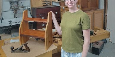 A woman proudly displays a wooden shelf she built in a woodworking shop.
