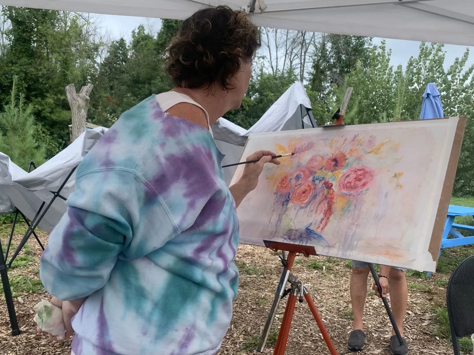 Person painting a floral artwork outdoors under a tent.