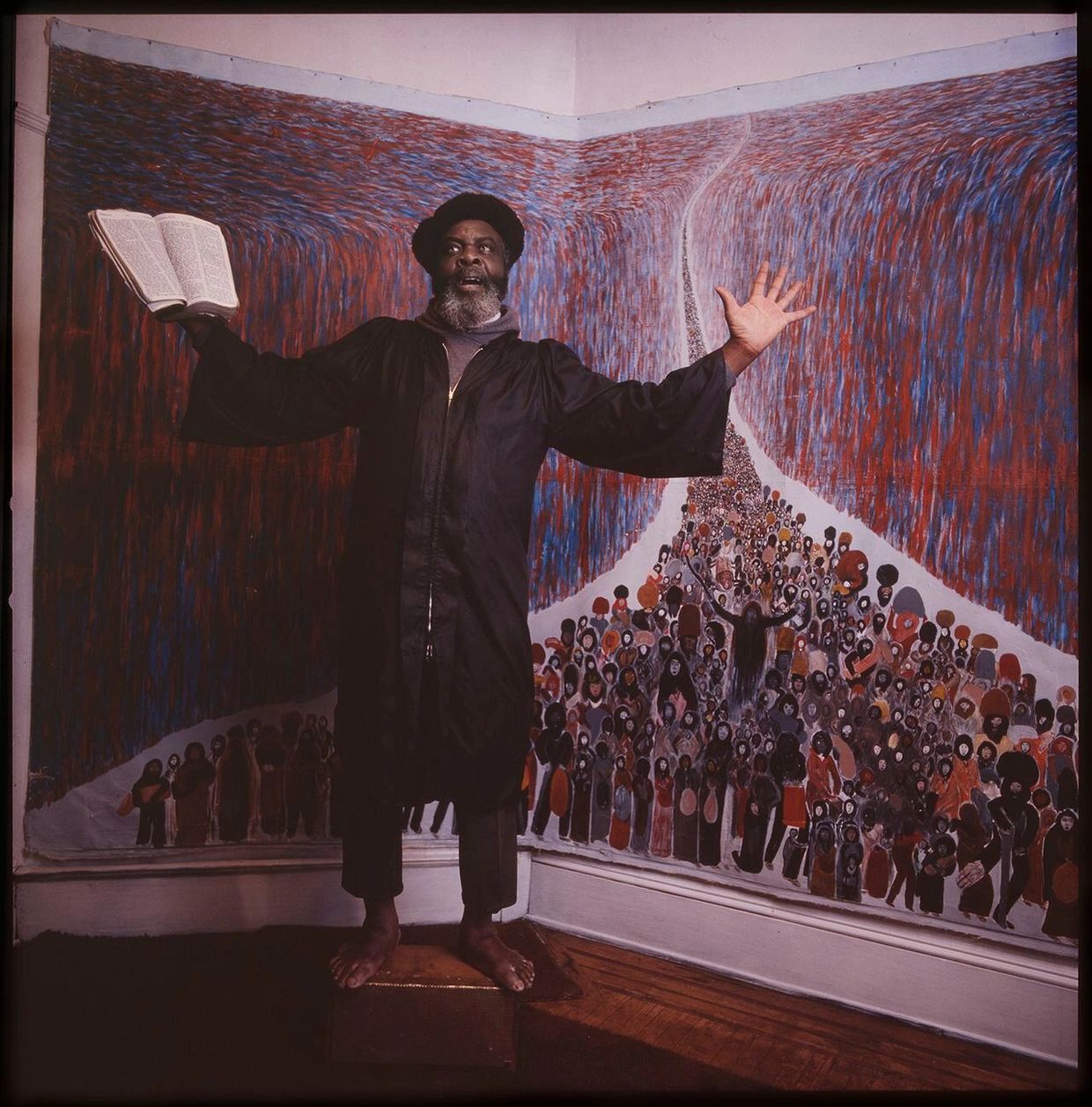 Man holding a book, standing barefoot on a box, in front of a vibrant mural.