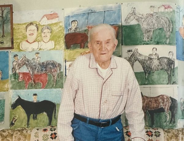 Elderly man stands in front of a wall covered with colorful drawings of horses and people.