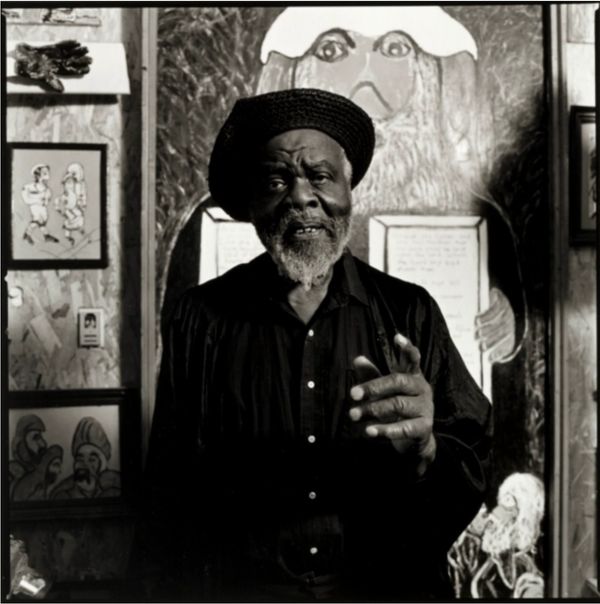 Black and white portrait of an elderly man with a beard and hat, standing in front of artwork.