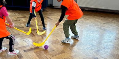 Children playing floor hockey indoors with orange vests and yellow sticks.