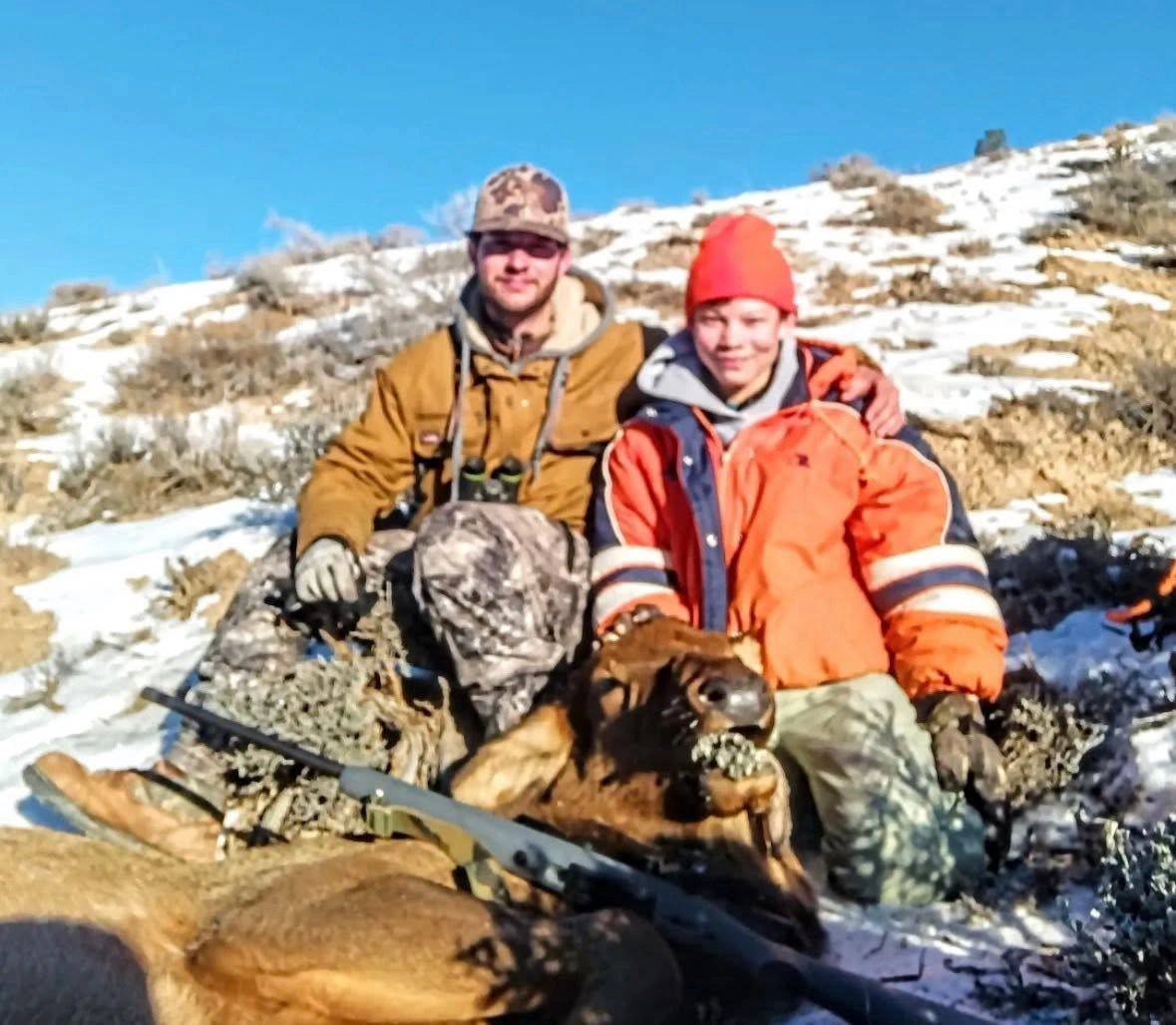 Two hunters pose with their kill on a snowy hillside under a clear blue sky.