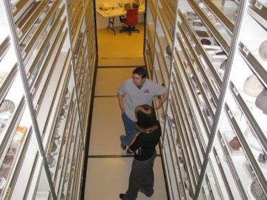 Two people talking between shelves of artifacts.