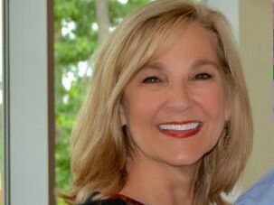 Smiling woman with blonde hair indoors near a window with greenery outside.