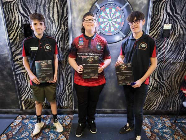 Three young dart players proudly holding certificates in front of a dartboard.