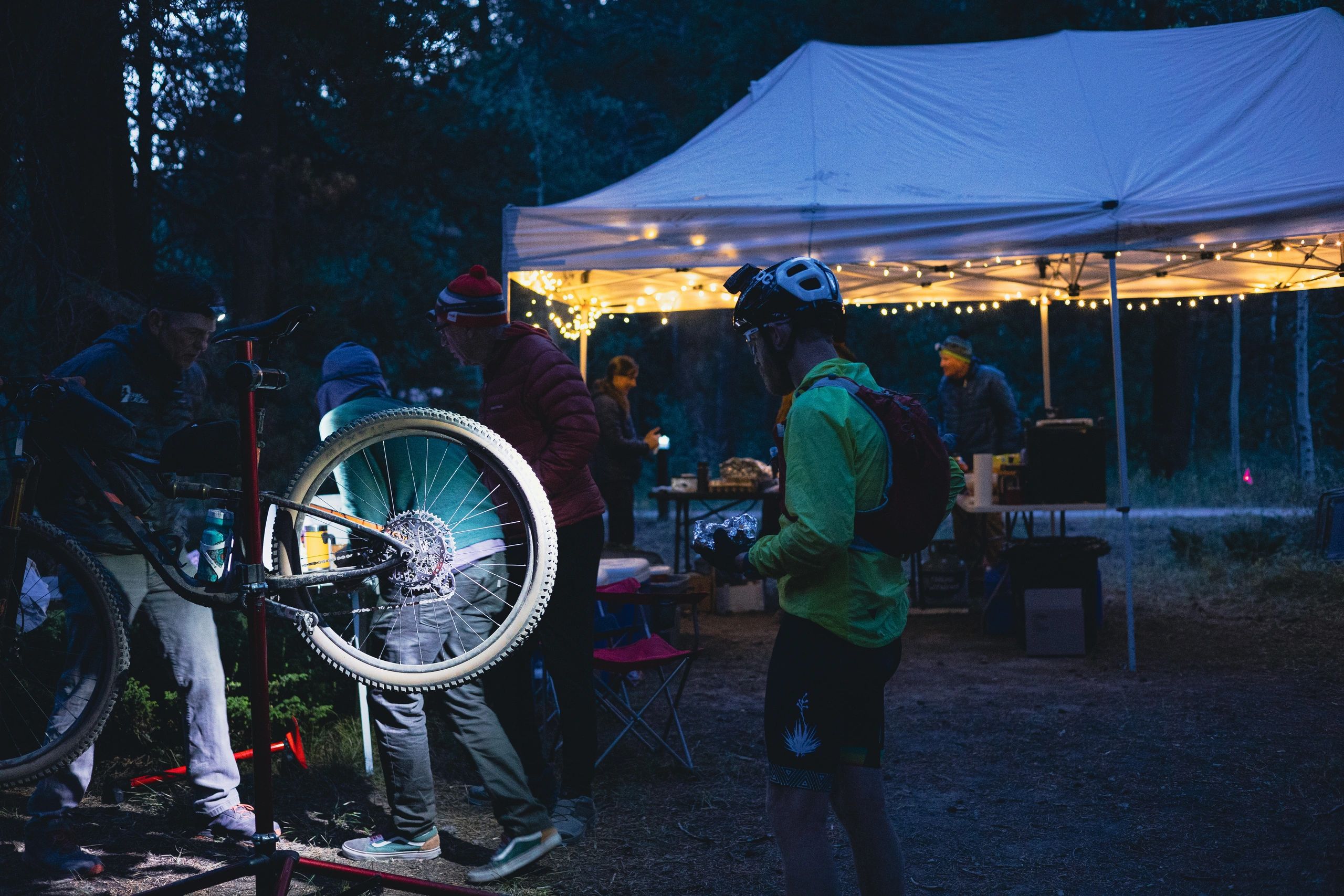 Cyclists gather at a lit tent in the forest during nighttime.