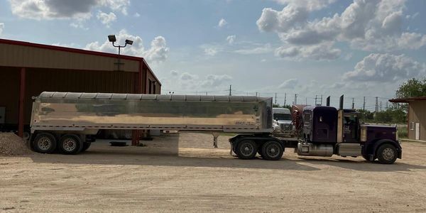A purple semi-truck with a shiny metal trailer parked on a dirt lot under a cloudy blue sky.