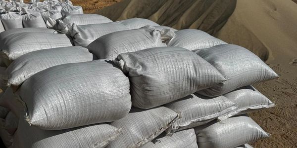 Stacked white sacks on a wooden pallet near sand piles under clear sky.