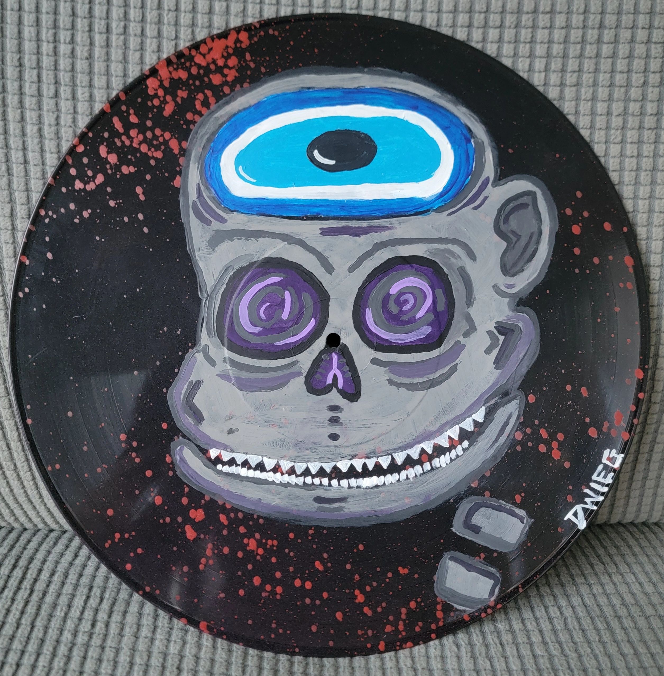 A painted skull with a blue eye on a black splattered background.