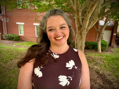 A smiling woman in a floral dress stands outdoors near trees and a brick building.