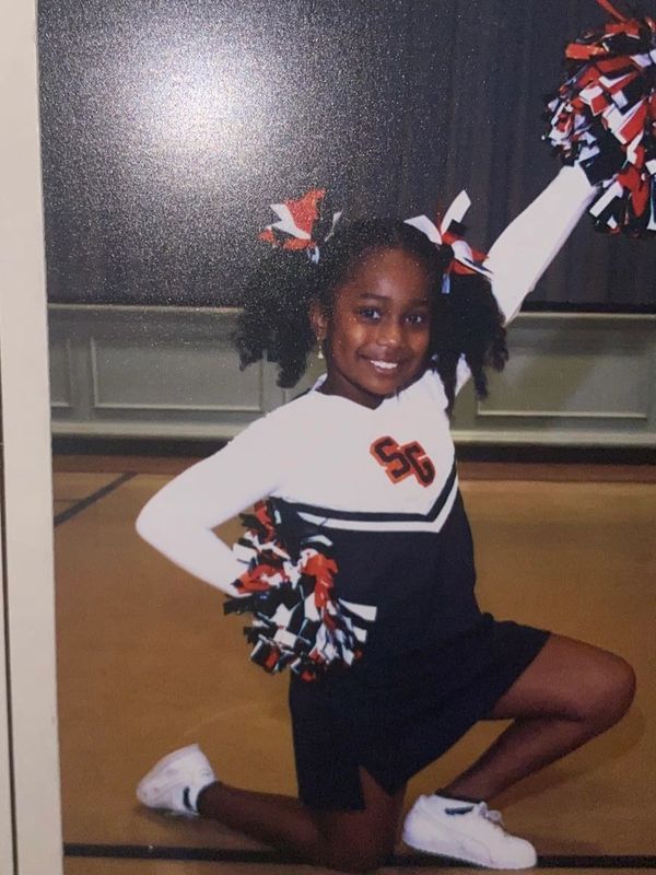 Young girl in cheerleading outfit kneeling with pom-poms, smiling.