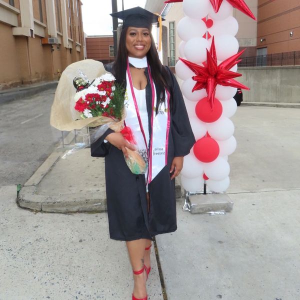 A smiling graduate in cap and gown holds flowers beside red and white balloon decorations.