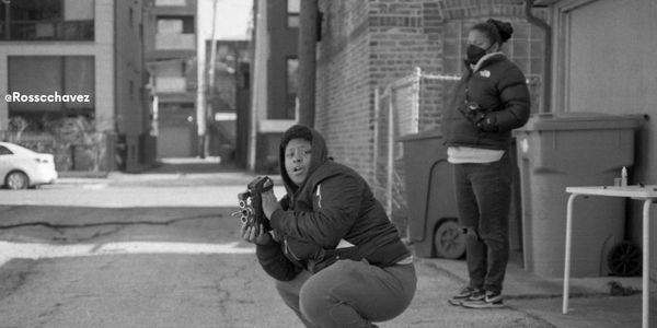 Two people in an urban alley, one crouching with a camera, the other standing wearing a mask.