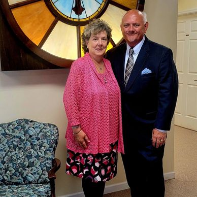 A couple posing indoors near a large clock with a floral chair beside them.
