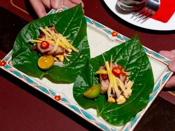 Hand holding a plate with two green leaf appetizers topped with meat, chili, and lime.