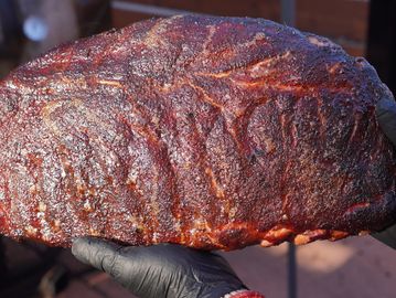 A gloved hand holding a large rack of smoked ribs with a dark, crispy bark.
