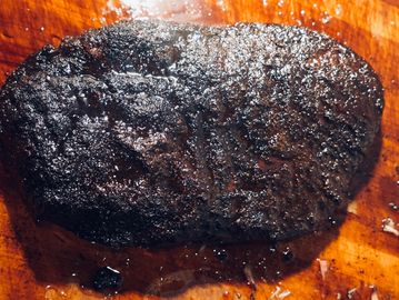 A charred, cooked steak resting on a wooden cutting board.