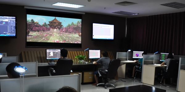 People working on computers in a dimly lit office with a large screen showing a scenic image.