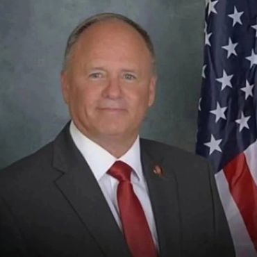 A man in a suit with a red tie standing beside an American flag.