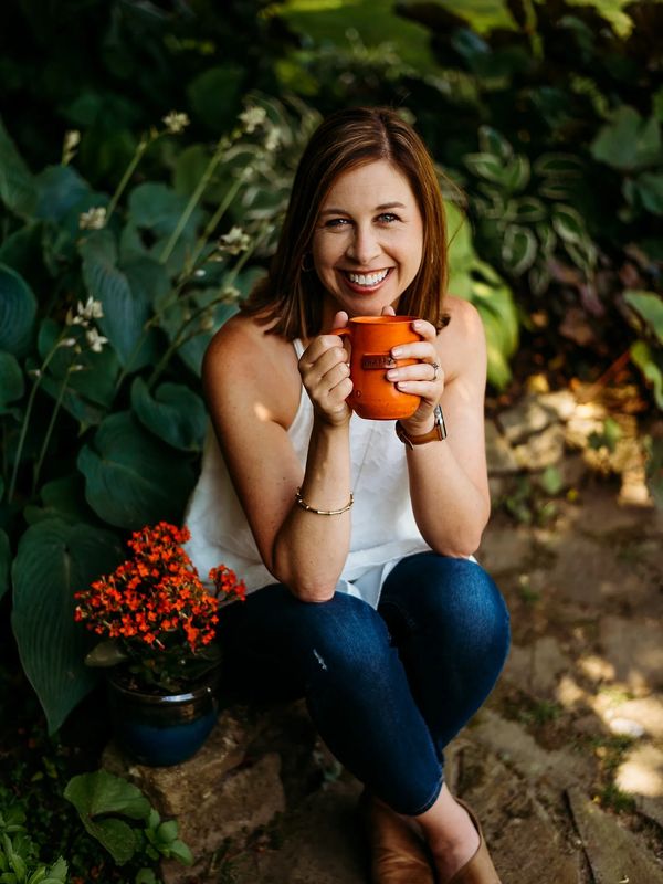 White female wearing a white shirt and jeans, with brown hair,  sitting outside in a rock garden hol