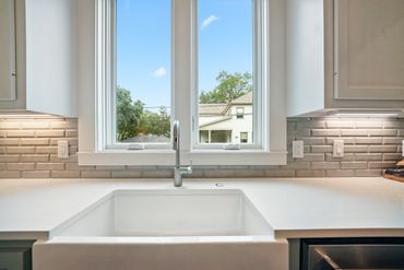 Modern kitchen sink under a large window with brick backsplash and white countertops.