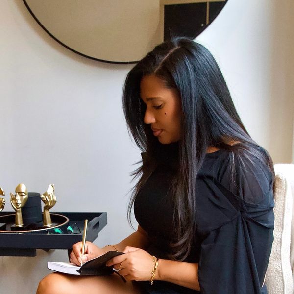 Woman in black dress writing in a notebook while seated on a cozy white chair.