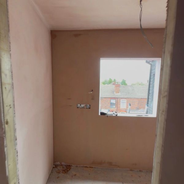 Small unfinished room with plastered walls and window showing houses outside.