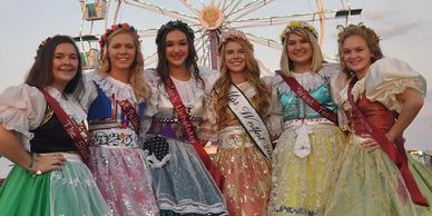 Six women in traditional dresses with sashes posing at a fairground.