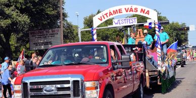 A red truck leads a festive parade with people waving and colorful decorations.
