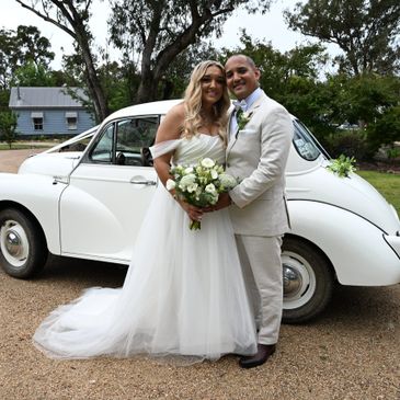 Newlywed couple posing happily in front of a vintage white car.