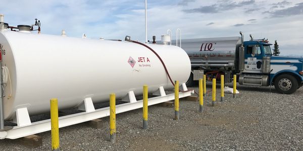 A large white Jet A fuel tank next to a blue tanker truck on gravel.