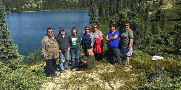 A group of people posing by a lake in a lush, forested area on a sunny day.