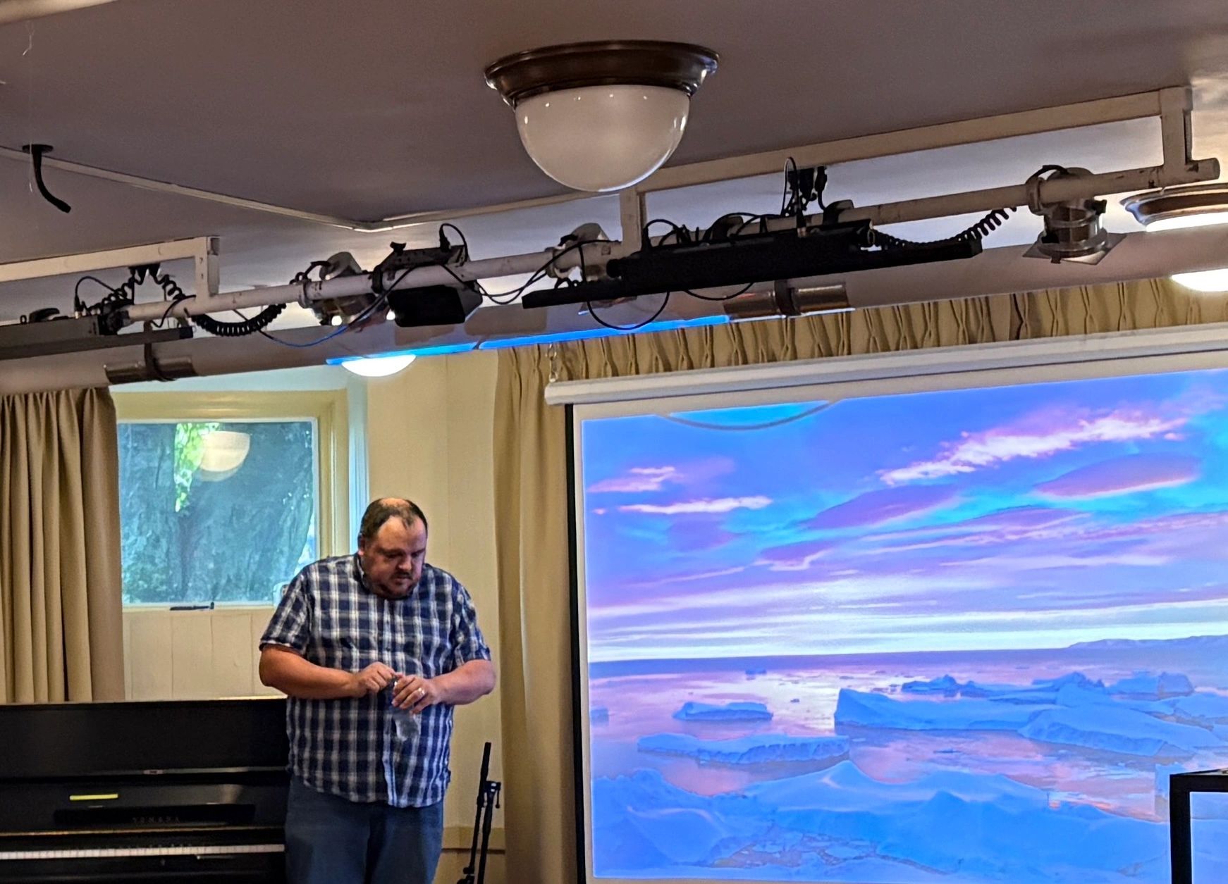 Man in plaid shirt standing near a piano and a large screen showing an icy landscape.