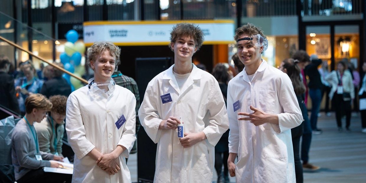 Three young men in lab coats at a science event, smiling and posing.